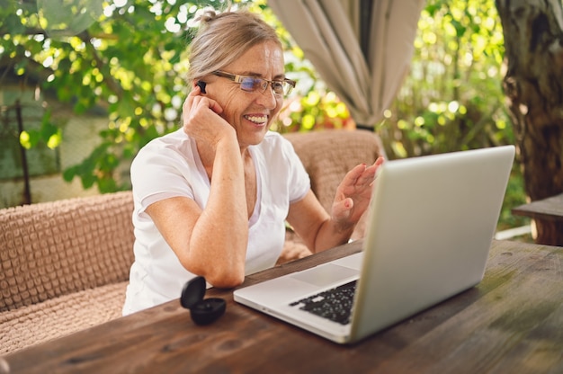 Foto technologie, alter leute konzept - ältere glückliche ältere frau, die drahtlose kopfhörer verwendet, die online mit laptop im freien im garten arbeiten. fernarbeit, fernunterricht.