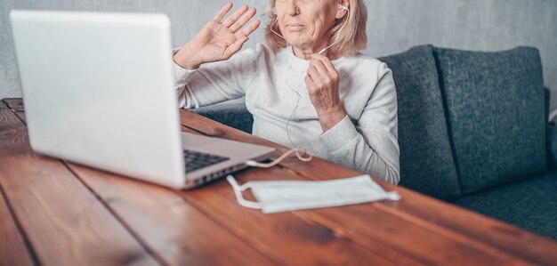 Technologie-, Alters- und Personenkonzept - glückliche ältere ältere Frau mit Gesichtsmaske, die arbeitet und einen Videoanruf mit Laptop-Computer zu Hause während der Coronavirus-COVID19-Pandemie macht. Zu Hause bleiben