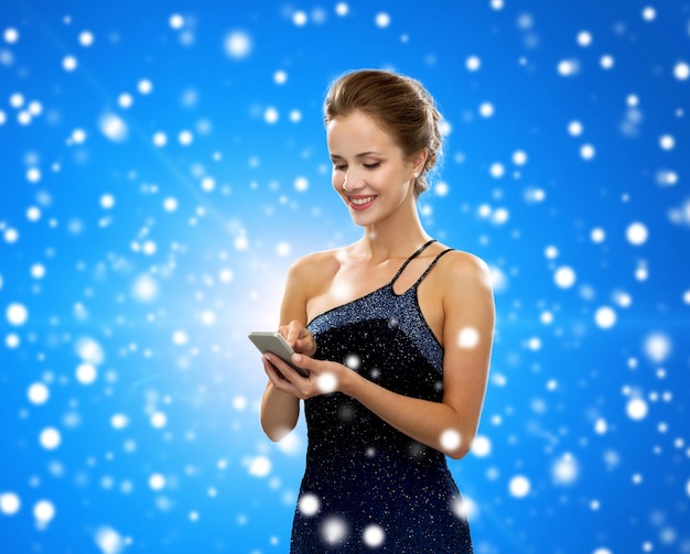 technologie, kommunikation, winterferien, weihnachten und personenkonzept - lächelnde frau im abendkleid, das smartphone über blauem schneebedecktem hintergrund hält