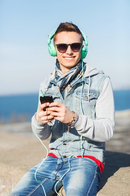 Technologie-, Lifestyle- und People-Konzept - lächelnder junger Mann oder Teenager mit Kopfhörern und Smartphone, der draußen Musik hört