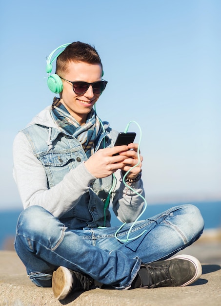 Technologie-, Lifestyle- und People-Konzept - lächelnder junger Mann oder Teenager mit Kopfhörern und Smartphone, der draußen Musik hört