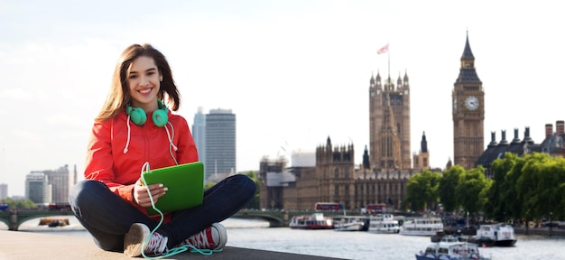 technologie-, reise-, tourismus-, musik- und personenkonzept - lächelnde junge frau oder jugendliches mädchen mit tablet-pc-computer und kopfhörern über london city und big ben tower hintergrund