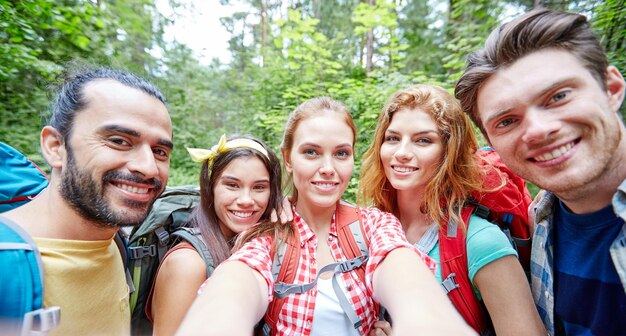 technologie-, reise-, tourismus-, wanderungs- und personenkonzept - gruppe lächelnder freunde, die mit rucksäcken spazieren gehen und selfie per smartphone oder kamera im wald machen