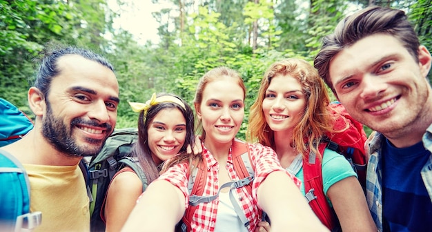 Foto technologie-, reise-, tourismus-, wanderungs- und personenkonzept - gruppe lächelnder freunde, die mit rucksäcken spazieren gehen und selfie per smartphone oder kamera im wald machen