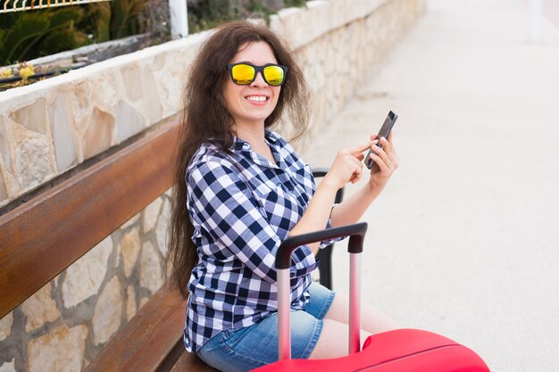 Technologie-, Reise- und Personenkonzept - junge Frau sitzt auf Bank in sonnigen Gläsern und tippt am Telefon.
