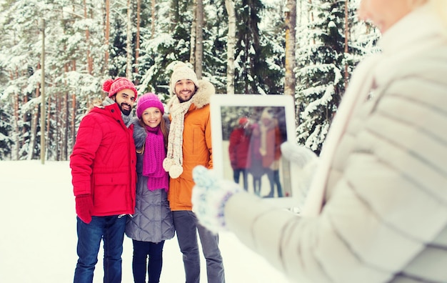 Technologie-, Saison-, Freundschafts- und Personenkonzept - Gruppe lächelnder Männer und Frauen, die mit Tablet-PC-Computer im Winterwald fotografieren