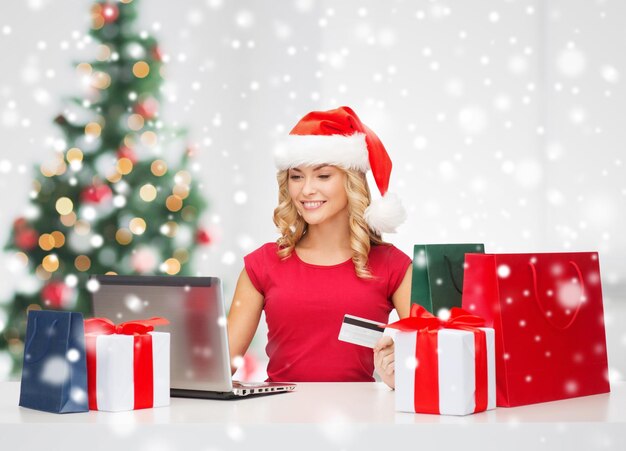 Technologie, Urlaub, Online-Shopping und People-Konzept - Frau in Weihnachtsmütze mit Geschenkboxen und Laptop-Computer über Wohnzimmerhintergrund