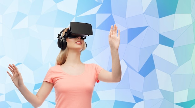 Technologie, virtuelle Realität, Unterhaltung und People-Konzept - glückliche junge Frau mit Virtual-Reality-Headset oder 3D-Brille und Kopfhörern, die ein Spiel über blauem Low-Poly-Texturhintergrund spielen