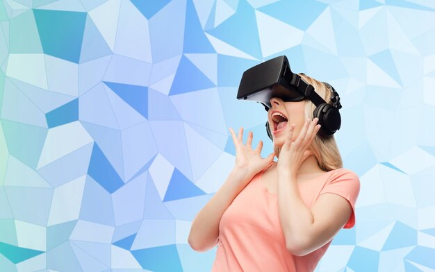 Foto technologie, virtuelle realität, unterhaltung und people-konzept - glückliche junge frau mit virtual-reality-headset oder 3d-brille und kopfhörern über blauem low-poly-texturhintergrund