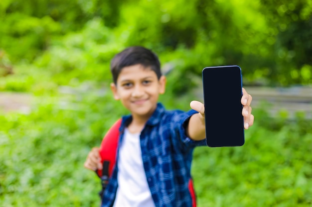 Technologiekonzept: Netter indischer kleiner Schuljunge, der Tasche hält und Smartphone zeigt