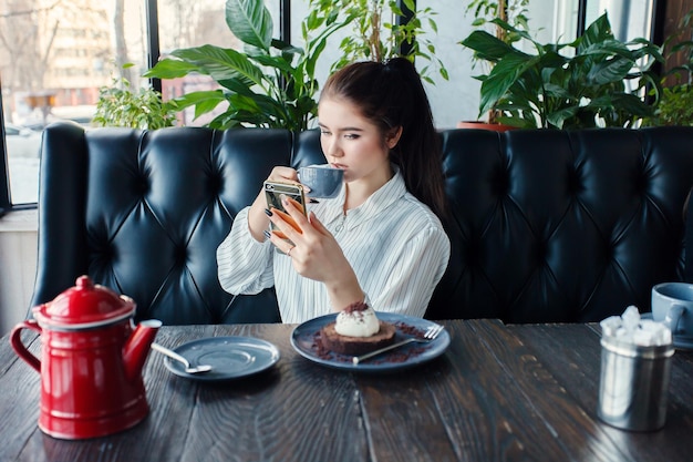 Technologien Emotionen Lifestyle Menschen Teenager-Konzept Junge glückliche weibliche Lesung auf ihrem Handy, während sie in einem modernen Café-Interieur sitzt Mädchen mit schönem Lächeln trinkt Tee