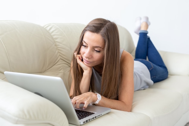 Technologien, Entspannung und freiberufliches Konzept - hübsche junge Frau, die auf Sofa liegt und am Laptop arbeitet