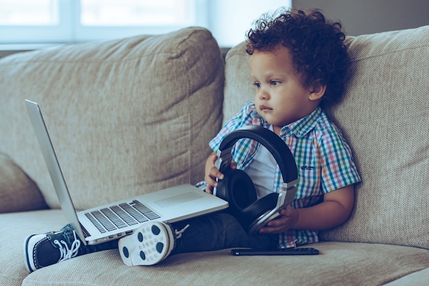 Technologien machen das Leben einfacher. Seitenansicht eines kleinen afrikanischen Jungen, der auf den Laptop schaut und Kopfhörer hält, während er zu Hause auf der Couch sitzt