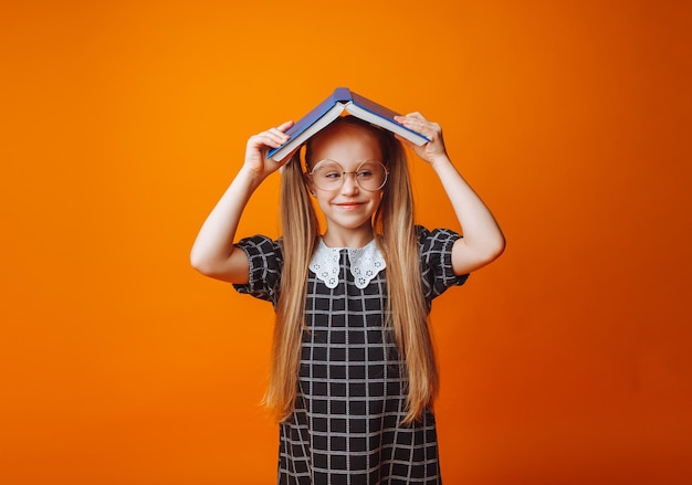 Un techo de libros sobre tu cabeza Una niña pequeña sostiene un libro de texto sobre su cabeza Una niña pequeña con un libro sobre un fondo naranja