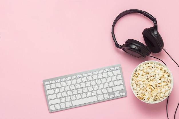 Teclado, auriculares y un tazón de palomitas de maíz sobre un fondo rosa. El concepto de ver películas, espectáculos, deportes en la PS, juegos en línea. Vista plana, vista superior.