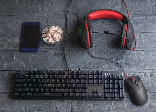 Teclado para jogos iluminado, fones de ouvido, mouse e marshmallow em uma mesa de madeira