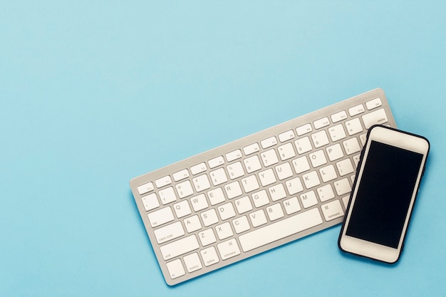 Teclado y teléfono móvil blanco sobre un fondo azul. Concepto de negocio, trabajo de oficina, aplicación móvil y sitio web. Vista plana, vista superior.