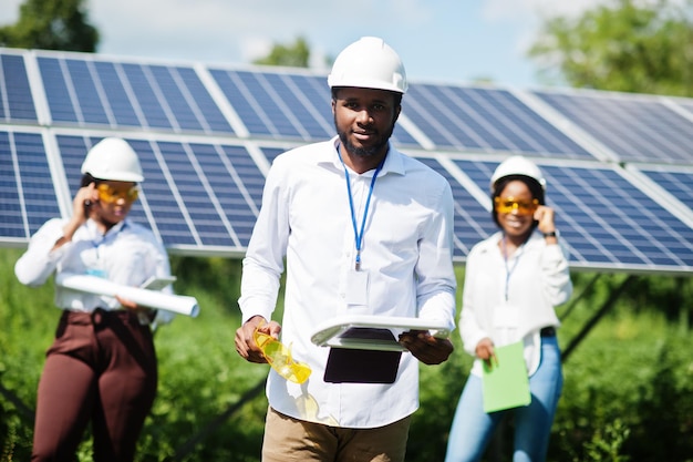 Técnico afro-americano verifica a manutenção dos painéis solares. Grupo de três engenheiros negros reunidos na estação solar.