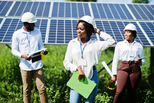 Técnico afroamericano comprueba el mantenimiento de los paneles solares. Grupo de tres ingenieros negros reunidos en la estación solar.