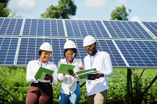 Técnico afroamericano comprueba el mantenimiento de los paneles solares. Grupo de tres ingenieros negros reunidos en la estación solar.