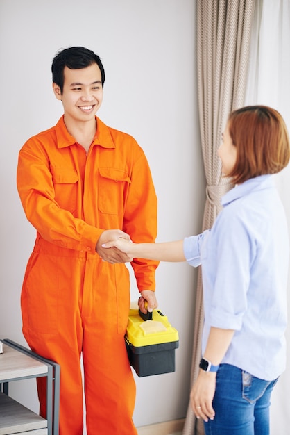 Técnico asiático sorridente com uniforme laranja e caixa de ferramentas apertando a mão da dona de casa