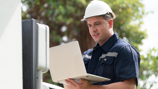 Técnico calificado que trabaja en la instalación de una estación de carga de vehículos eléctricos en el hogar, solucionando problemas y configurando el sistema de carga con una computadora portátil para vehículos eléctricos en casa Synchronos