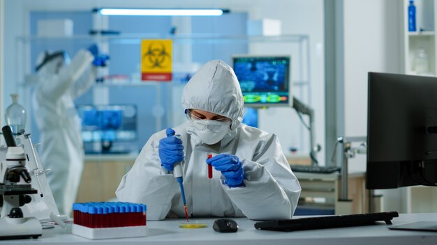 Técnico científico en traje de ppe con micropipeta y placa de Petri trabajando en un laboratorio de investigación analizando muestras de sangre. Equipo que examina la evolución del virus para el desarrollo de vacunas contra covid19