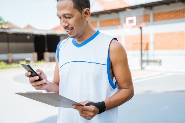 Foto técnico de basquete olhando para a tela de um telefone celular enquanto segura uma prancheta