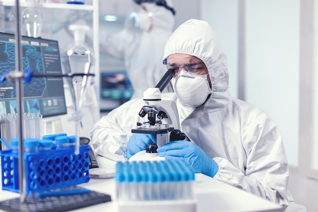 Técnico de laboratorio en equipo de ppe examinando muestras de virus bajo microscopio en laboratorio. Científico en traje de protección sentado en el lugar de trabajo utilizando tecnología médica moderna durante la epidemia global.