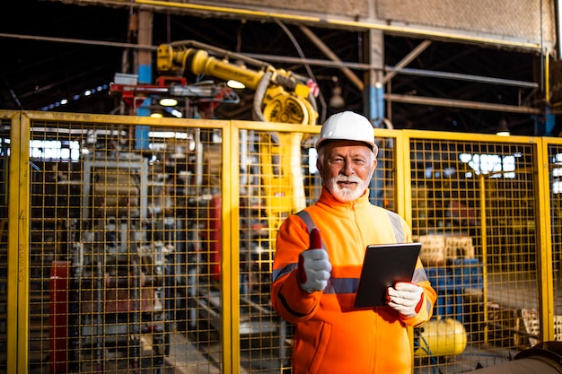 técnico de máquinas industriales sosteniendo los pulgares hacia arriba frente al brazo robótico en la fábrica de fabricación