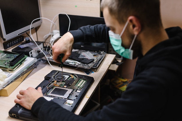 Foto técnico reparando una computadora portátil en el laboratorio. concepto de reparación informática, electrónica, actualización, tecnología. coronavirus. hombre trabajando, con máscara protectora en taller.