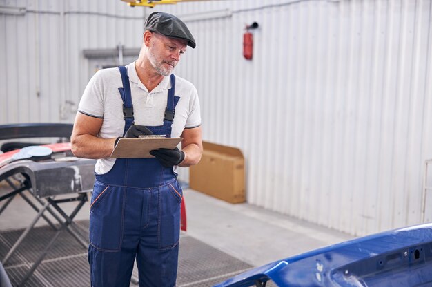 Técnico de sexo masculino barbudo en monos de trabajo mirando automóvil y sonriendo mientras escribe en el portapapeles