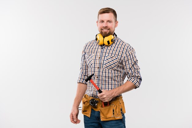 Foto técnico sonriente barbudo con auriculares protectores, martillo y cinturón de herramientas de pie frente a la cámara de forma aislada