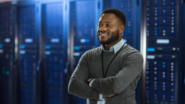 Técnico de TI de centro de datos negro de pie en medio de un corredor de rack de servidores está de pie con los brazos cruzados y está sonriendo