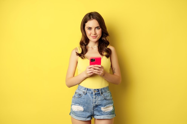 Tecnología celular y concepto de compras en línea. Mujer joven con estilo usando teléfono inteligente, aplicación de teléfono móvil, de pie sobre fondo amarillo.