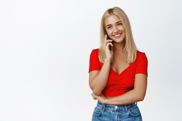 Tecnología celular Sonriente mujer rubia hablando por teléfono móvil haciendo una llamada de pie sobre fondo blanco.