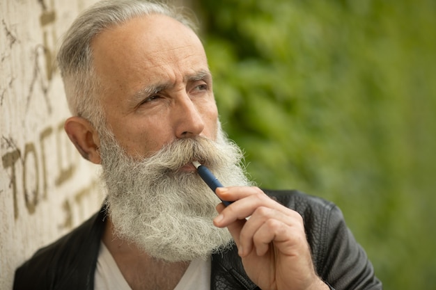 Tecnología de cigarrillos electrónicos. Sistema IQOS de tabaco. Primer plano de un hombre mayor barbudo fumando un cigarrillo híbrido eléctrico con una almohadilla térmica.