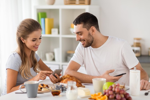 Foto tecnología comer y conceptos de personas pareja feliz con teléfonos inteligentes desayunando en casa