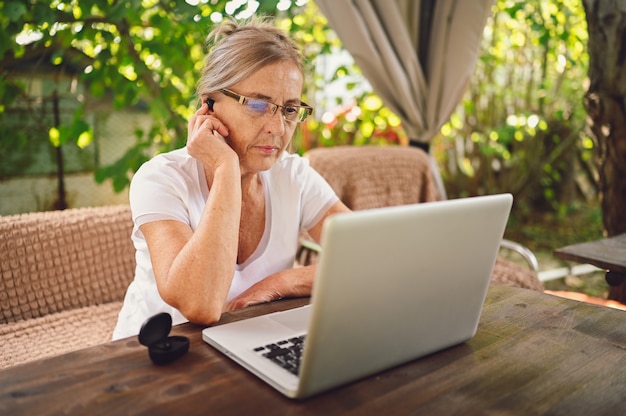 Tecnologia, conceito de pessoas de idade avançada - mulher idosa feliz sênior usando fones de ouvido sem fio trabalhando online com o computador laptop ao ar livre no jardim. trabalho remoto, educação a distância.