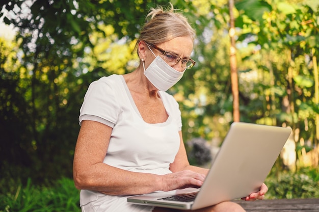 Tecnologia, conceito de pessoas de idade avançada - mulher idosa sênior na máscara protetora facial trabalhando online com o computador portátil ao ar livre no jardim. Trabalho remoto, educação a distância.