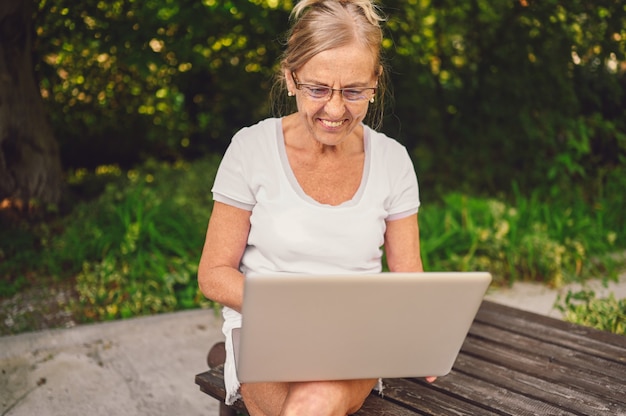 Tecnología, concepto de personas de la vejez - anciana mayor feliz trabajando en línea con la computadora portátil al aire libre en el jardín. Trabajo a distancia, educación a distancia.