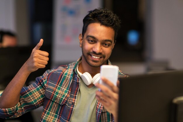 Foto tecnologia de comunicação e conceito de prazo homem criativo com smartphone tendo chamada de vídeo no escritório à noite e mostrando polegar para cima