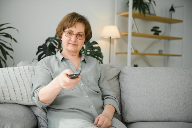 Tecnologia de televisão idade e conceito de pessoas feliz mulher sênior assistindo tv e mudando de canal com controle remoto em casa