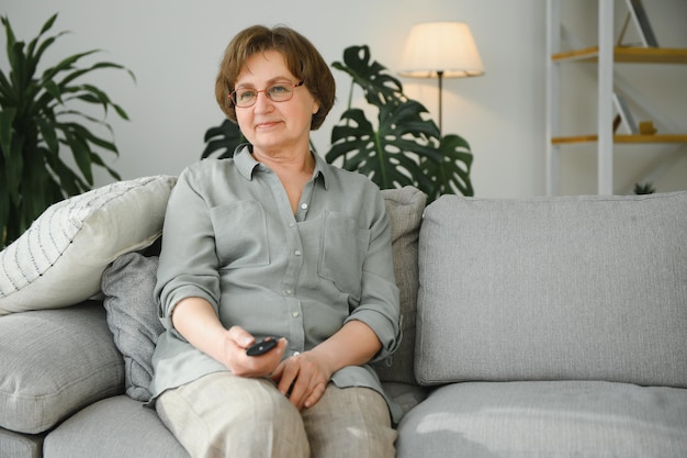 Tecnologia de televisão idade e conceito de pessoas feliz mulher sênior assistindo tv e mudando de canal com controle remoto em casa