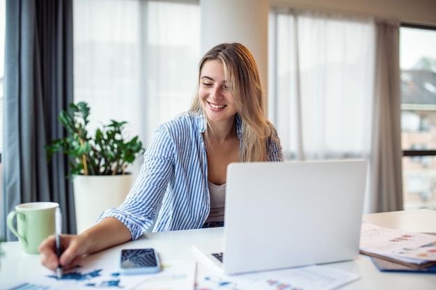 Tecnologia de trabalho remoto e conceito de pessoas feliz sorridente jovem empresária com computador portátil e papéis trabalhando em home office