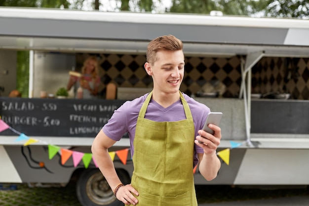 Foto tecnologia de venda de rua e conceito de pessoas jovem vendedor feliz em avental com smartphone em caminhão de comida