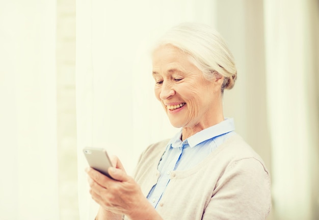 tecnología, edad de comunicación y concepto de personas - mujer mayor feliz con mensaje de texto de teléfono inteligente en casa