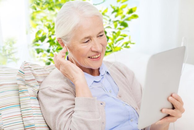 tecnología, edad y concepto de personas - mujer anciana feliz con tableta, computadora y auriculares escuchando música en casa sobre un fondo natural verde