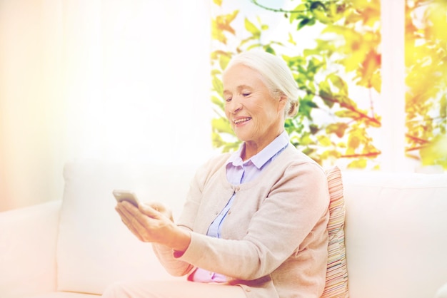 tecnología, edad y concepto de personas - mujer anciana feliz con teléfono inteligente mensaje de texto en casa sobre la ventana con fondo natural verde