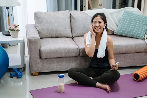 Tecnologia esportiva e conceito de estilo de vida saudável. mulher chinesa asiática sorridente em fones de ouvido sem fio, ouvindo música no smartphone na sala de estar de casa. jovem fitness em fones de ouvido bluetooth.